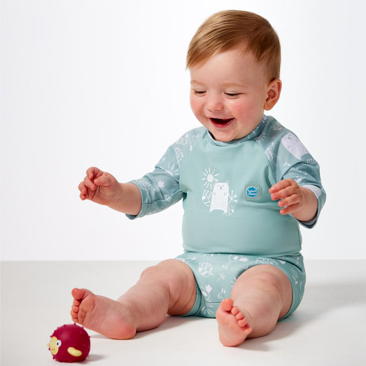 Lifestyle image of child wearing Happy Nappy Sunsuit in greenish blue and white, and animals themed print including bears, birds and llamas. Front.