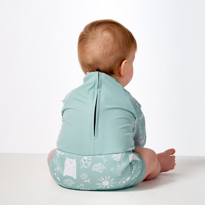 Lifestyle image of child wearing Happy Nappy Sunsuit in greenish blue and white, and animals themed print including bears, birds and llamas. Back.