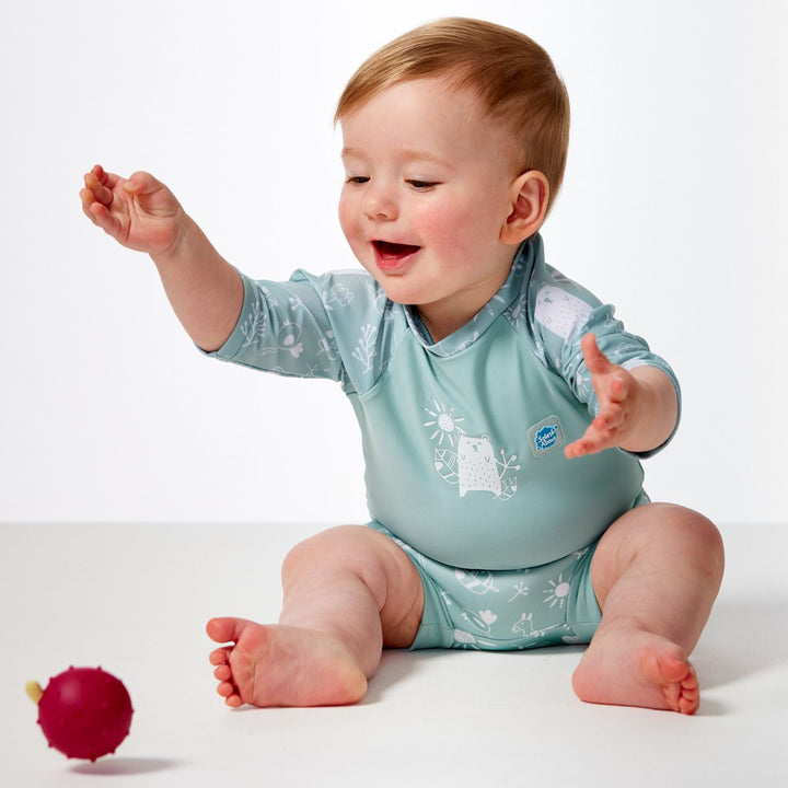 Lifestyle image of child wearing Happy Nappy Sunsuit in greenish blue and white, and animals themed print including bears, birds and llamas. Front.