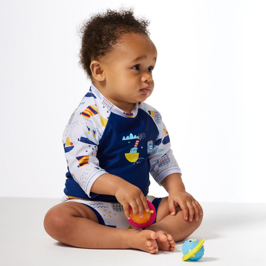 Lifestyle image of child wearing Happy Nappy Sunsuit in white and navy, and boats themed print. Side.