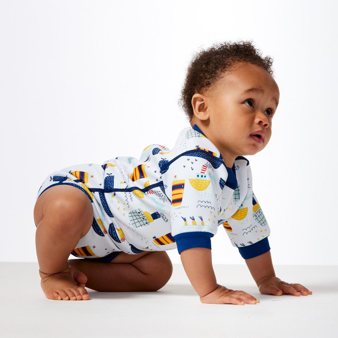 Lifestyle image of child wearing a baby wetsuit with built in swim nappy in white with navy trims and boats themed print. Side.