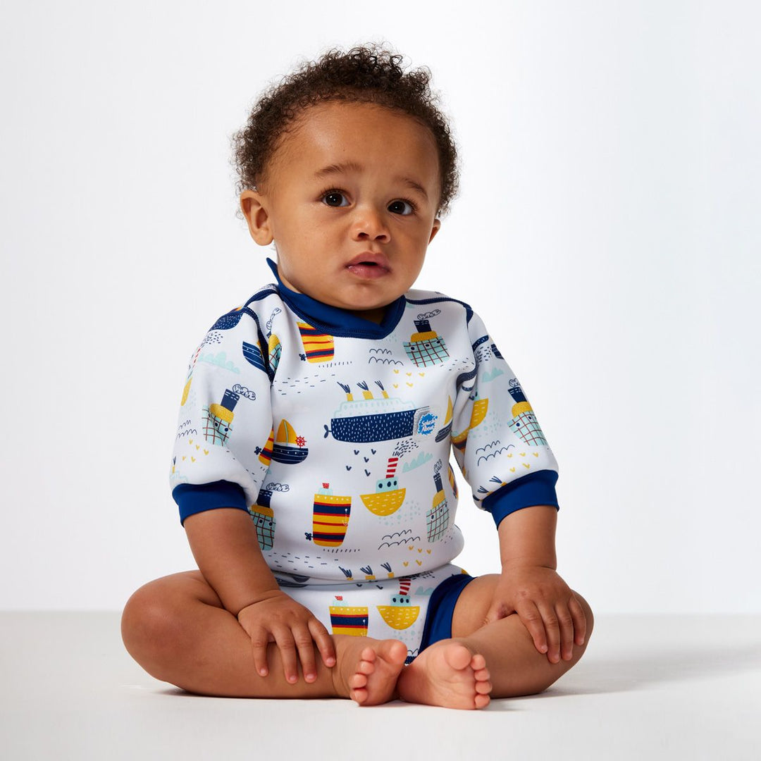 Lifestyle image of child wearing a baby wetsuit with built in swim nappy in white with navy trims and boats themed print. Front.