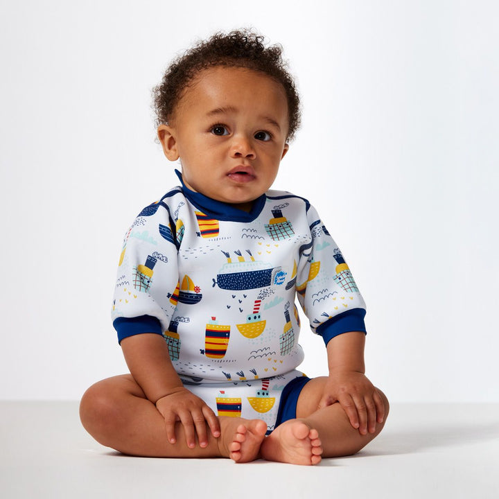 Lifestyle image of child wearing a baby wetsuit with built in swim nappy in white with navy trims and boats themed print. Front.