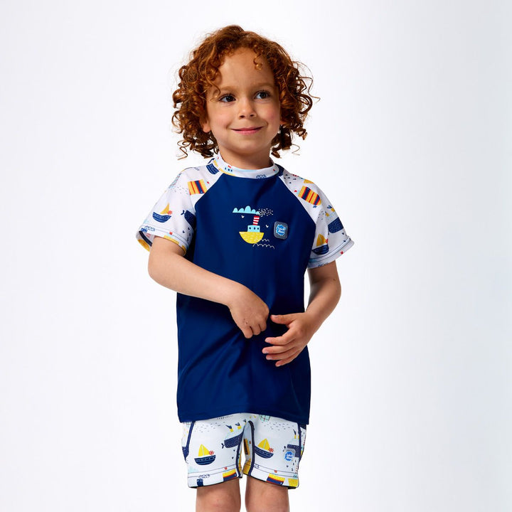 Lifestyle image of child wearing UV protective short sleeve rash top in blue, and boats themed print on the chest and sleeves. He's also wearing matching jammers. Front.
