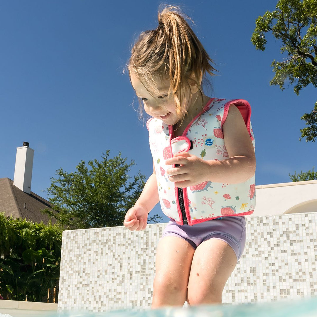 Lifestyle image of child wearing neoprene swim vest for toddlers with non-removable floats in white, pink trims and forest themed print, including hedgehogs, mushrooms and leaves. Front.