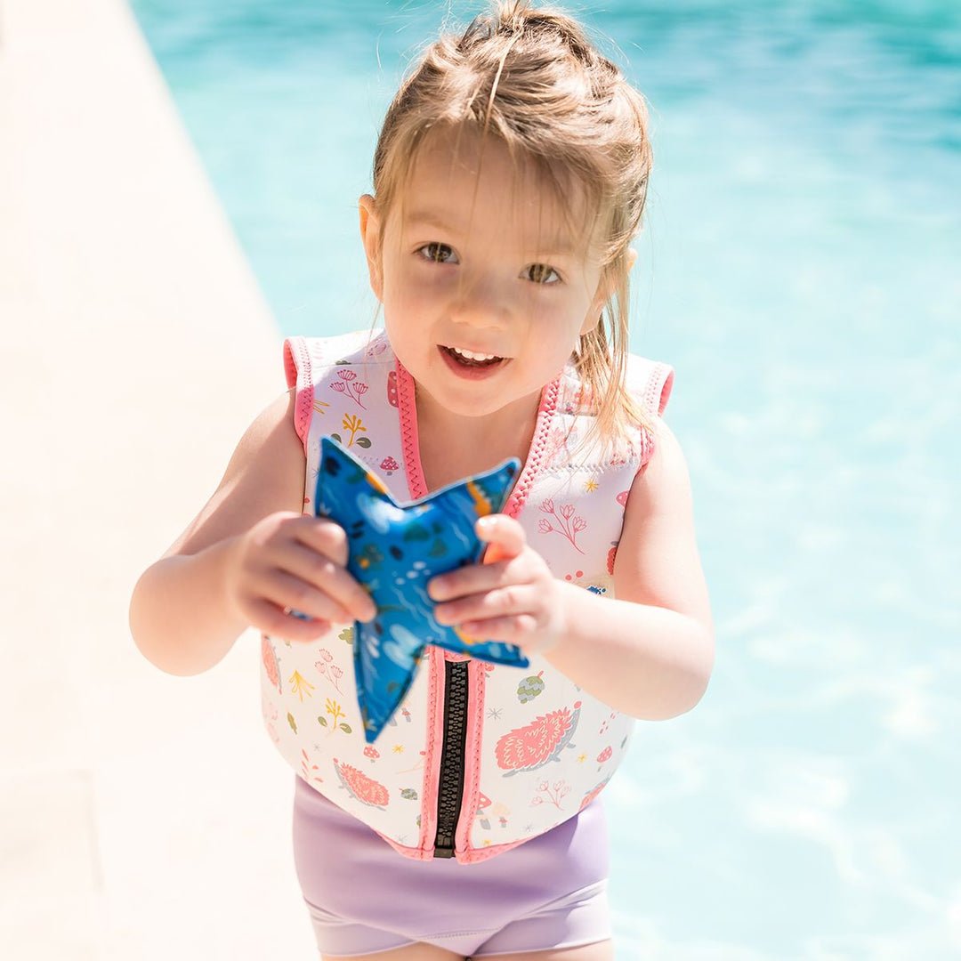 Lifestyle image of child wearing neoprene swim vest for toddlers with non-removable floats in white, pink trims and forest themed print, including hedgehogs, mushrooms and leaves. Front.