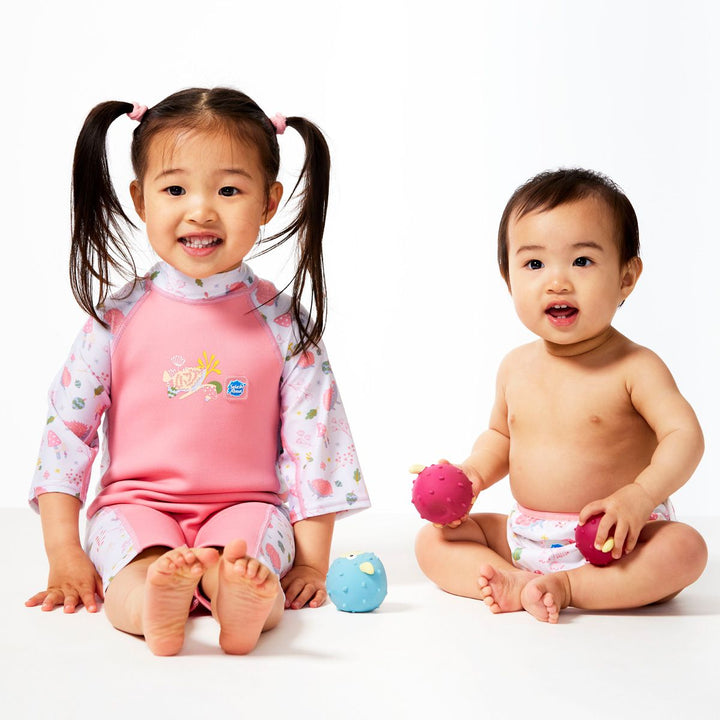 Lifestyle image of child wearing one piece UV sun and sea wetsuit for toddlers in pink. Forest themed print including hedgehogs, mushrooms and leaves on sleeves, side panels, neck and chest. Front. Baby sitting side by side, wearing matching nappy wrap.