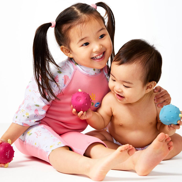 Lifestyle image of child wearing one piece UV sun and sea wetsuit for toddlers in pink. Forest themed print including hedgehogs, mushrooms and leaves on sleeves, side panels, neck and chest. Front. Baby sitting side by side, wearing matching nappy wrap. They're playing with pufferfish toys.