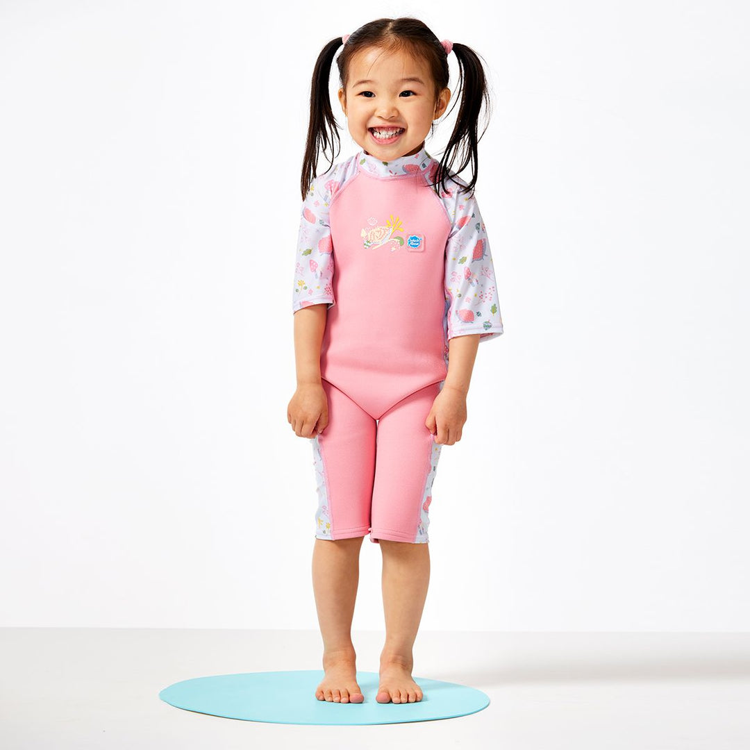 Lifestyle image of child wearing one piece UV sun and sea wetsuit for toddlers in pink. Forest themed print including hedgehogs, mushrooms and leaves on sleeves, side panels, neck and chest. Front.