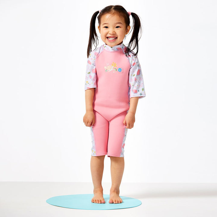 Lifestyle image of child wearing one piece UV sun and sea wetsuit for toddlers in pink. Forest themed print including hedgehogs, mushrooms and leaves on sleeves, side panels, neck and chest. Front.
