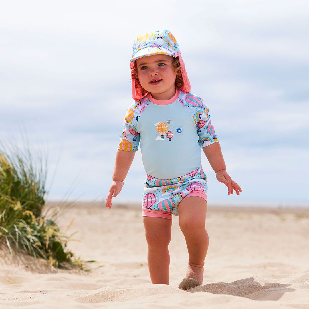 Lifestyle image of child wearing a Happy Nappy Sunsuit in baby blue with pink trims and hot air balloons themed print, including rainbows and clouds. Front. She's also wearing a matching legionnaire hat.