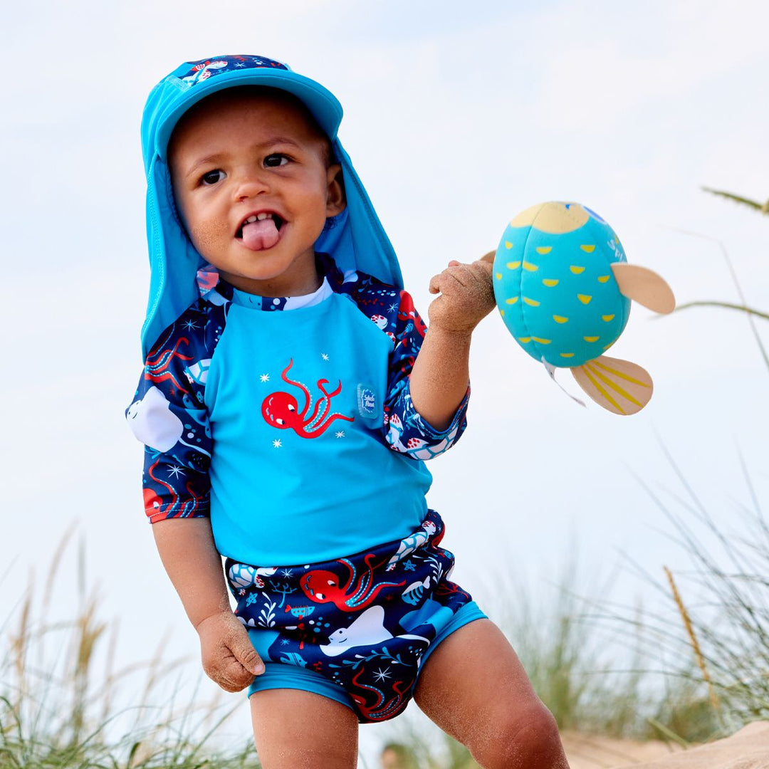 Lifestyle image of toddler wearing a Happy Nappy Sunsuit in navy blue and cyan, with under the sea themed print, including turtles, stingrays, octopus, fish and more. Front. He's also wearing matching legionnaire hat.