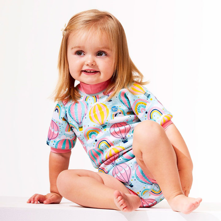 Lifestyle image of toddler wearing a cute wetsuit with built in swim nappy in baby blue with pink trims and hot air balloons themed print, including rainbows and clouds.