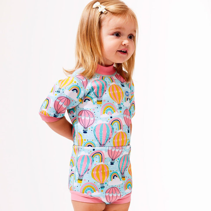 Lifestyle image of toddler wearing a cute wetsuit with built in swim nappy in baby blue with pink trims and hot air balloons themed print, including rainbows and clouds.