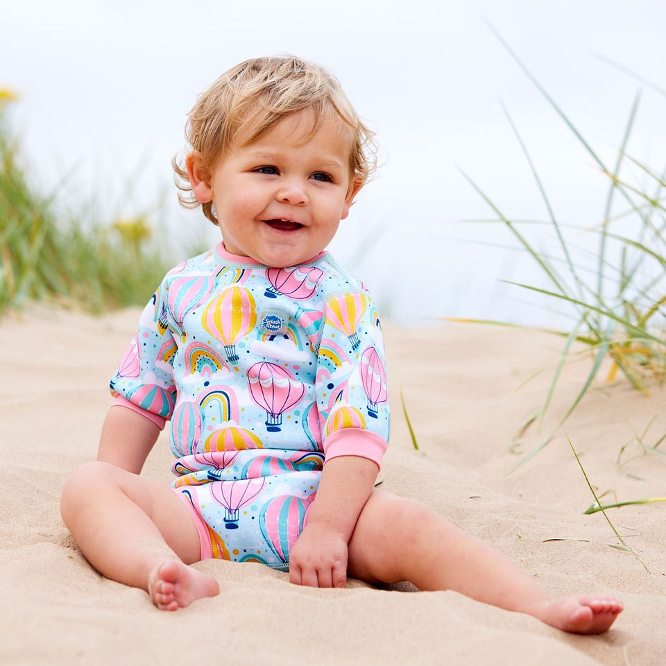 Lifestyle image of baby wearing a cute wetsuit with built in swim nappy in baby blue with pink trims and hot air balloons themed print, including rainbows and clouds.