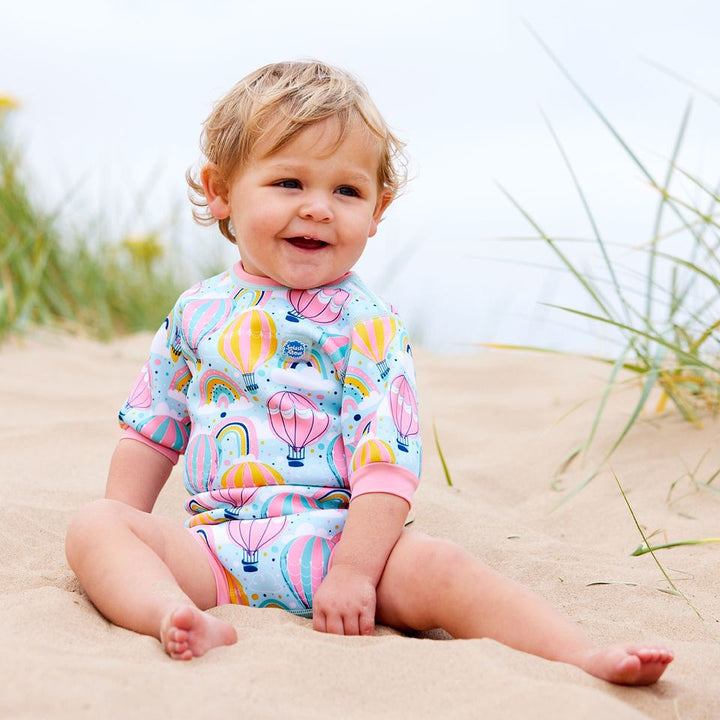 Lifestyle image of baby wearing a cute wetsuit with built in swim nappy in baby blue with pink trims and hot air balloons themed print, including rainbows and clouds.