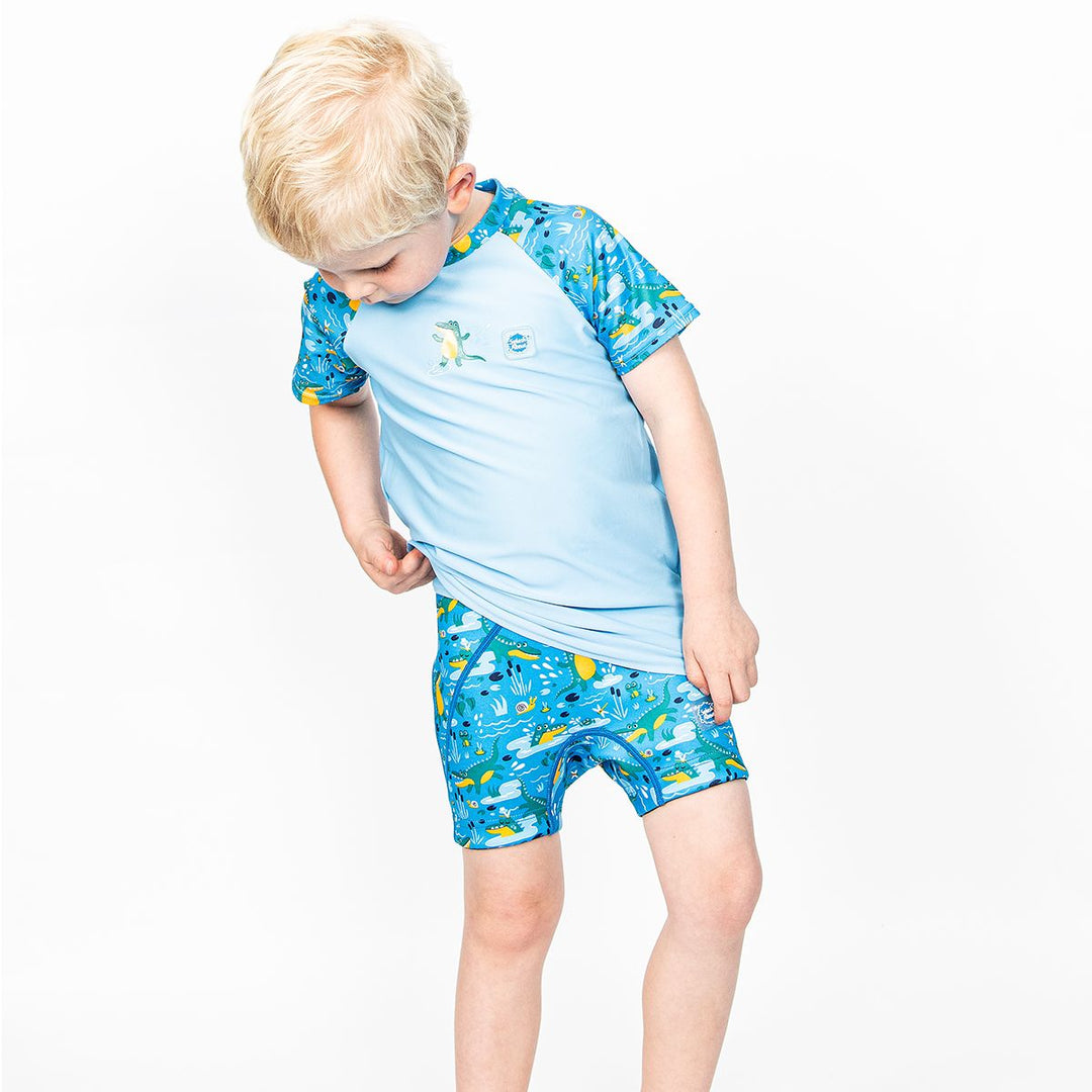 Lifestyle image of toddler sitting by the pool, wearing neoprene swim shorts in blue with light blue waist and swamp themed print, including crocodiles, frogs, snails and more.