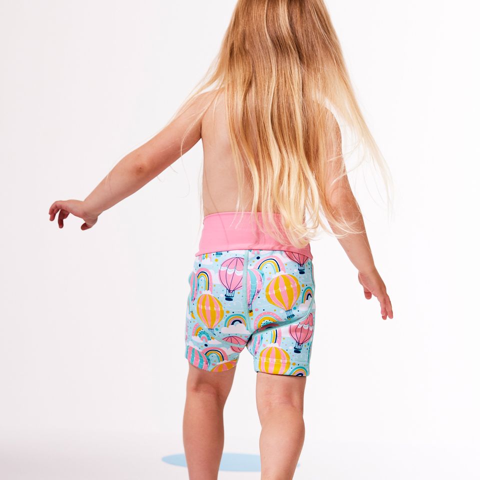 Lifestyle image of child wearing neoprene swim shorts in baby blue with pink waist and hot air balloons themed print, including clouds and rainbows.