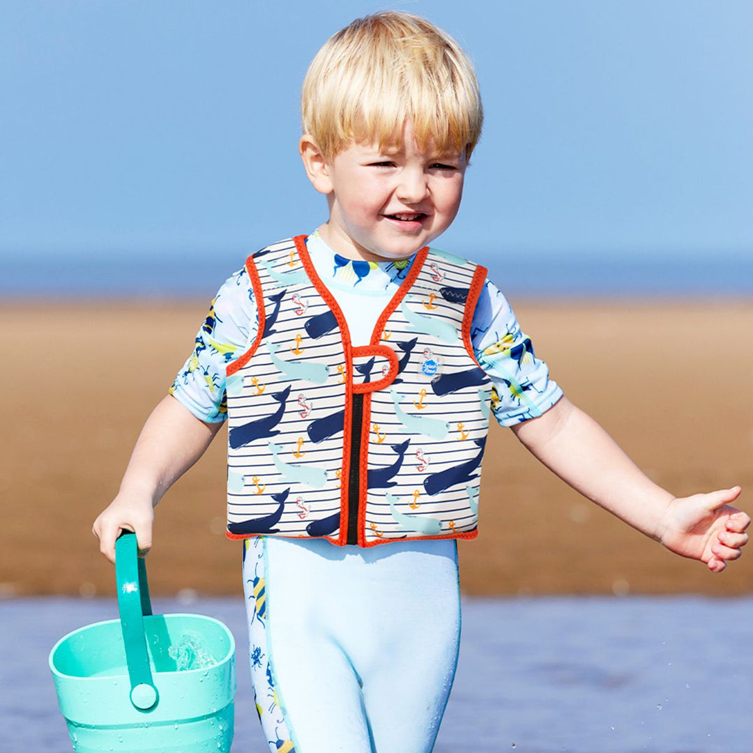 Lifestyle image of child wearing neoprene swim vest for toddlers with non-removable floats in white with red trims, whales themed print and navy blue stripes. 