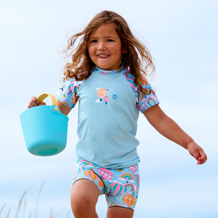 Lifestyle image of child wearing neoprene swim shorts in baby blue with pink waist and hot air balloons themed print, including clouds and rainbows.
