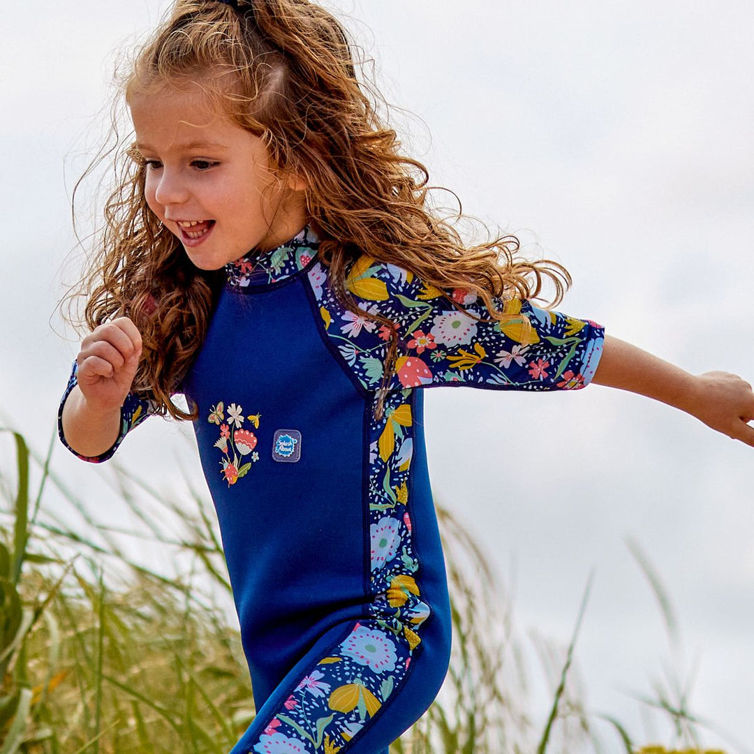 Lifestyle image of child wearing a one piece UV sun and sea wetsuit for toddlers in navy blue with yellow trims. Flowers and insects themed print on sleeves, side panels and neck.