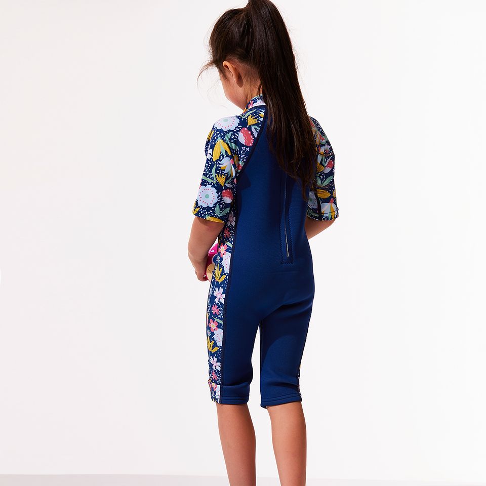 Lifestyle image of child wearing a one piece UV sun and sea wetsuit for toddlers in navy blue with yellow trims. Flowers and insects themed print on sleeves, side panels and neck.
