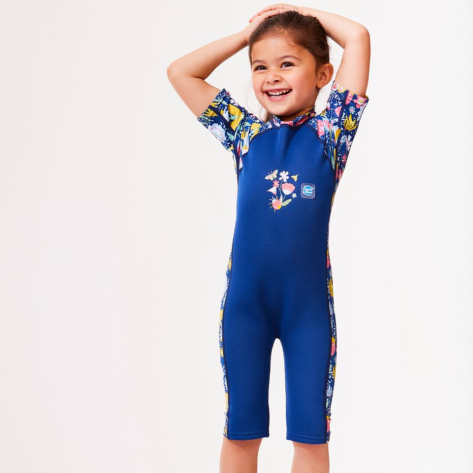 Lifestyle image of child wearing a one piece UV sun and sea wetsuit for toddlers in navy blue with yellow trims. Flowers and insects themed print on sleeves, side panels and neck.
