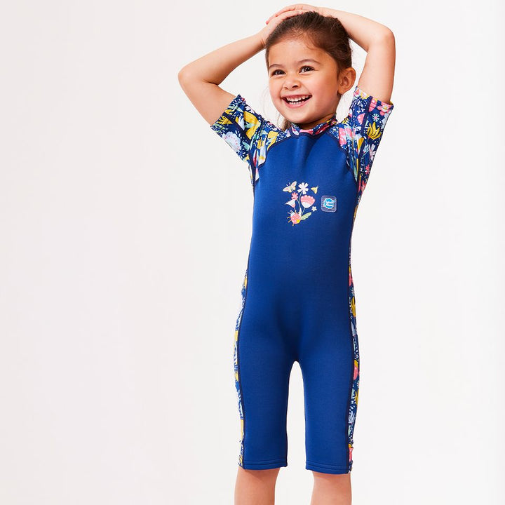 Lifestyle image of child wearing a one piece UV sun and sea wetsuit for toddlers in navy blue with yellow trims. Flowers and insects themed print on sleeves, side panels and neck.