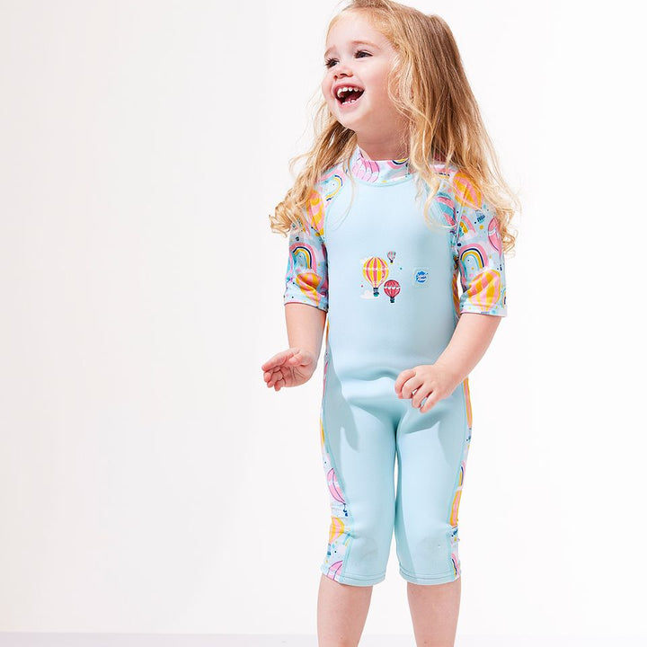 Lifestyle image of a child wearing a one piece UV sun and sea wetsuit for toddlers in light blue. Hot air balloons themed print including clouds and rainbows on sleeves, side panels, neck and chest.