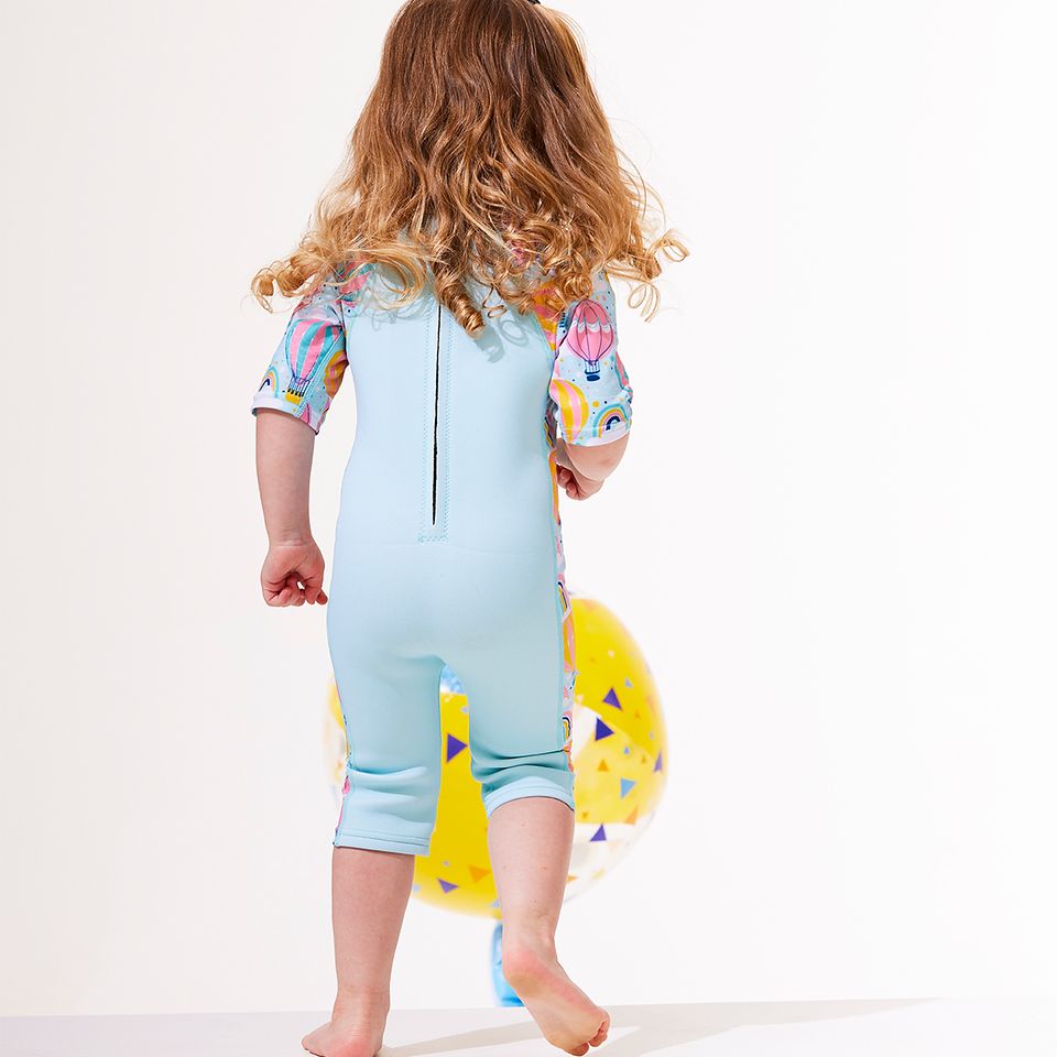Lifestyle image of a child wearing a one piece UV sun and sea wetsuit for toddlers in light blue. Hot air balloons themed print including clouds and rainbows on sleeves, side panels, neck and chest.