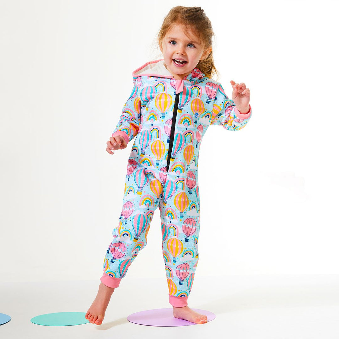 Lifestyle image of toddler wearing a waterproof fleece-lined onesie with hood in baby blue and hot air balloons themed print, including rainbows and clouds. Pink trims.