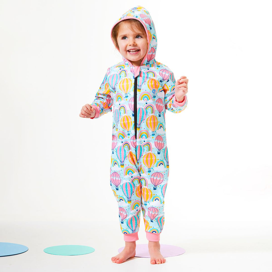 Lifestyle image of toddler wearing a waterproof fleece-lined onesie with hood in baby blue and hot air balloons themed print, including rainbows and clouds. Pink trims.