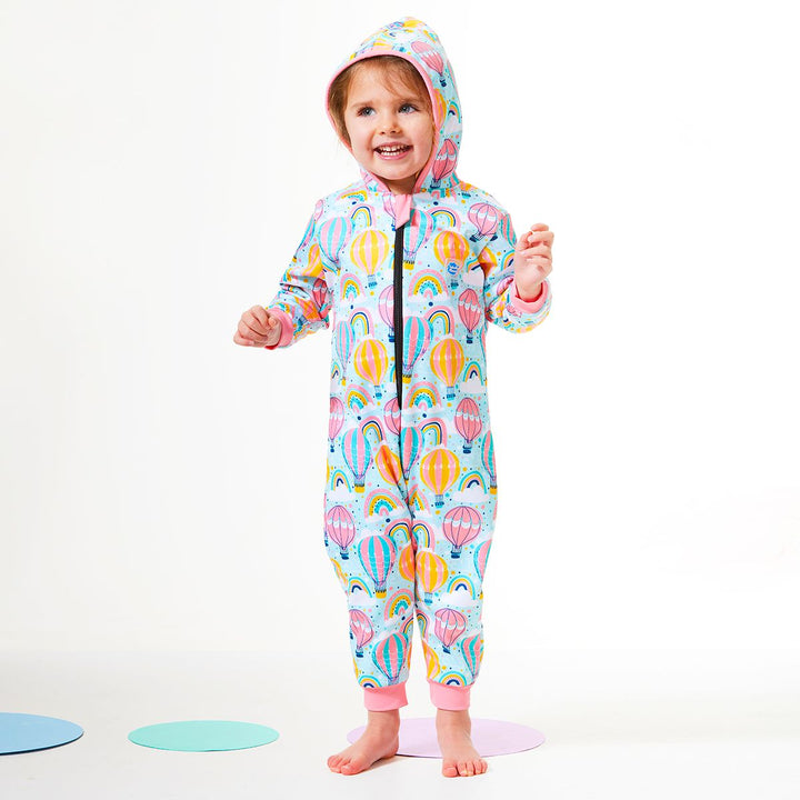 Lifestyle image of toddler wearing a waterproof fleece-lined onesie with hood in baby blue and hot air balloons themed print, including rainbows and clouds. Pink trims.