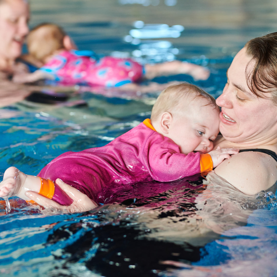 Baby girl wearing pink and orange Warm in One wet suit during Water Babies swim lesson. The carer is holding her in the water.