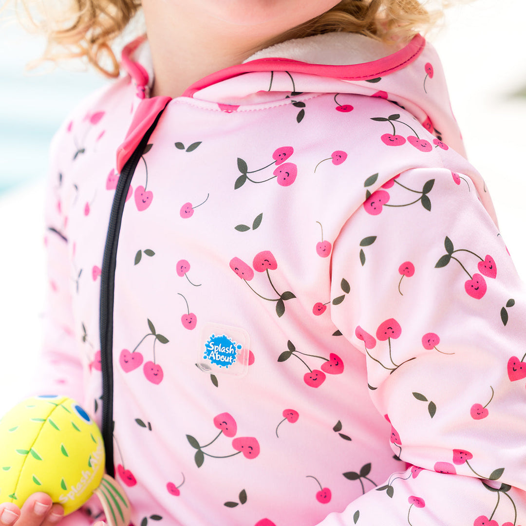Close up showing little one wearing waterproof fleece-lined onesie with hood in pink and cherries themed print. Hot pink trims.