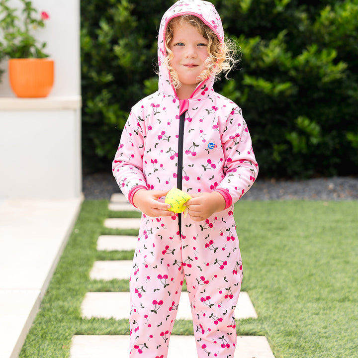 Little one wearing waterproof fleece-lined onesie with hood in pink and cherries themed print. Hot pink trims. Front.