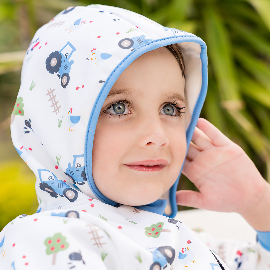 Little one wearing waterproof fleece-lined onesie with hood in white and farm themed print. Blue trims.