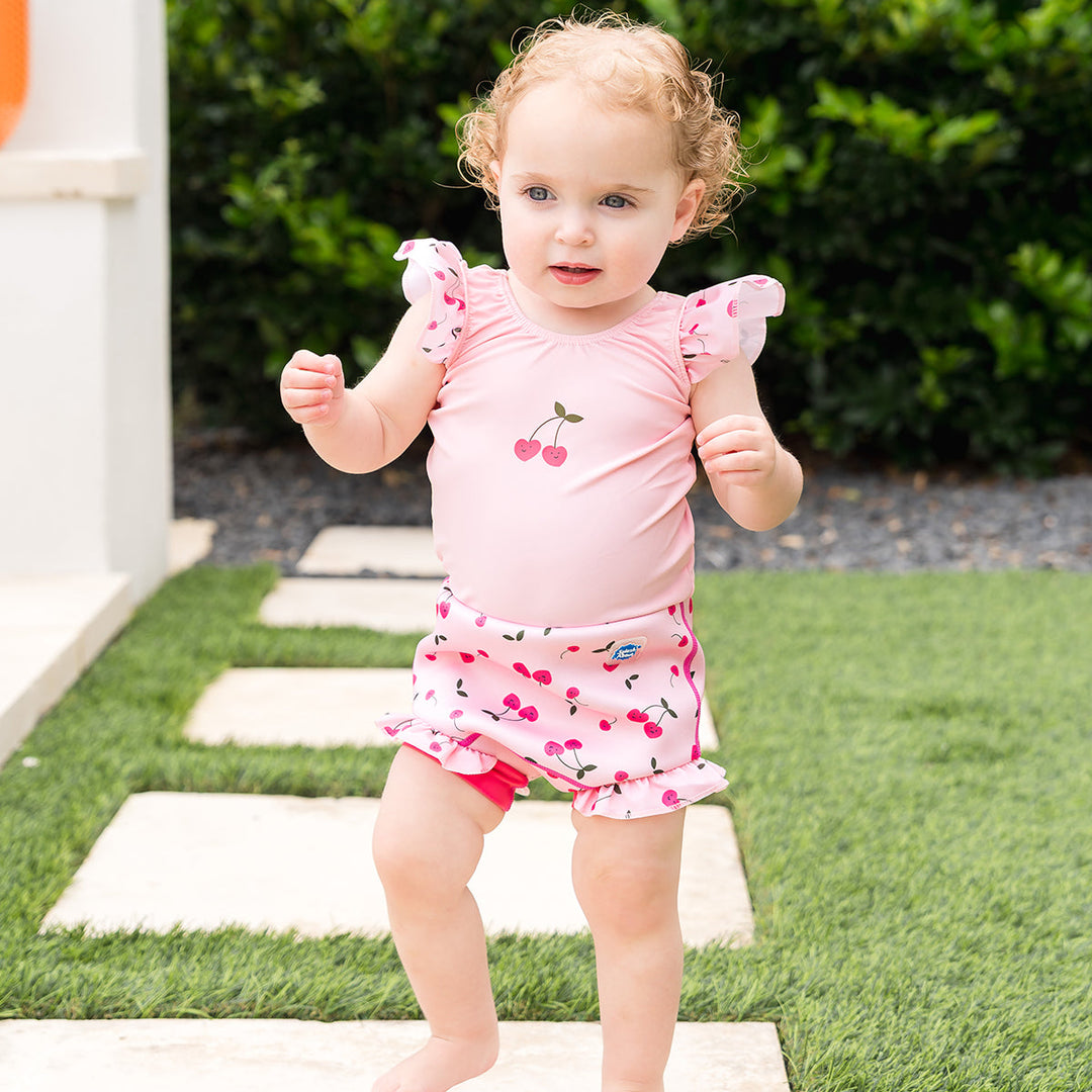 Little one wearing Happy Nappy Costume in pink and cherries themed print, featuring frills in arms and legs.