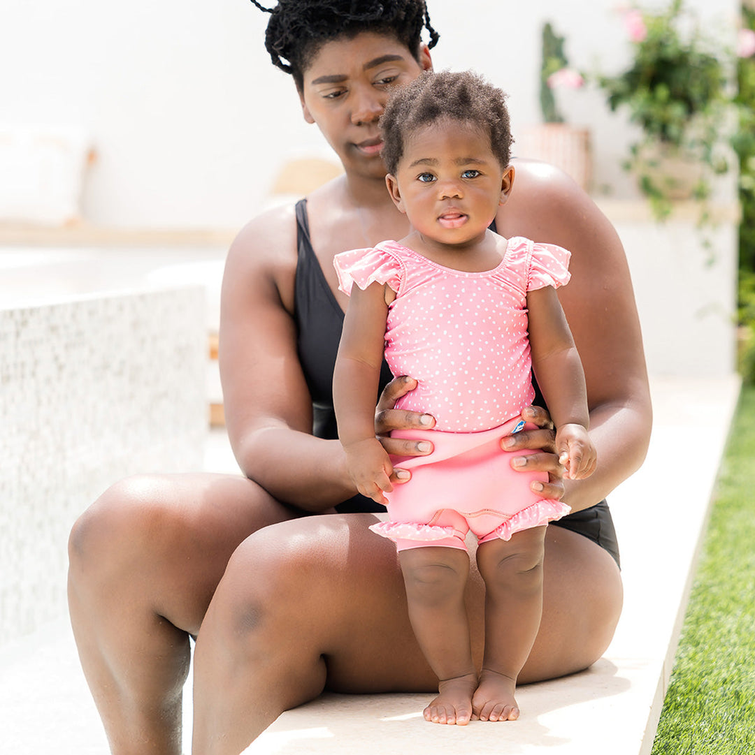 Little one wearing Happy Nappy Costume in pink and white dots, featuring frills in arms and legs, poolside with mum.