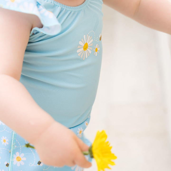 Little one wearing Happy Nappy Costume in light blue and daisies and bees themed print, featuring frills in arms and legs.