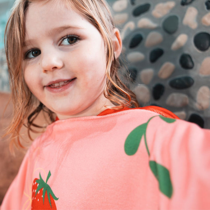 Little one wearing hooded poncho in red and pink with strawberries themed print.
