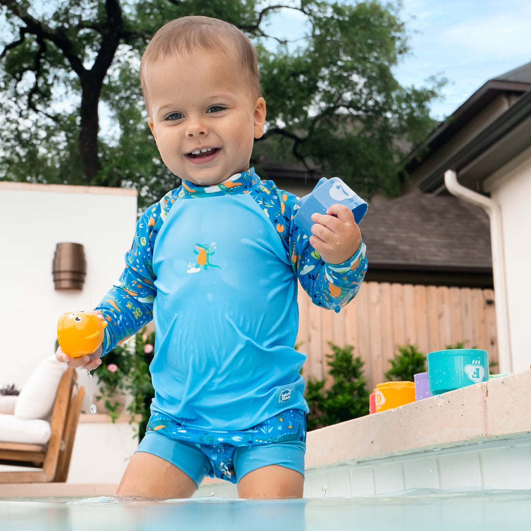 Little one wearing UV protective long sleeve rash top in blue, featuring crocodiles and frogs themed print on the chest and sleeves. They are also wearing matching Happy Nappy.