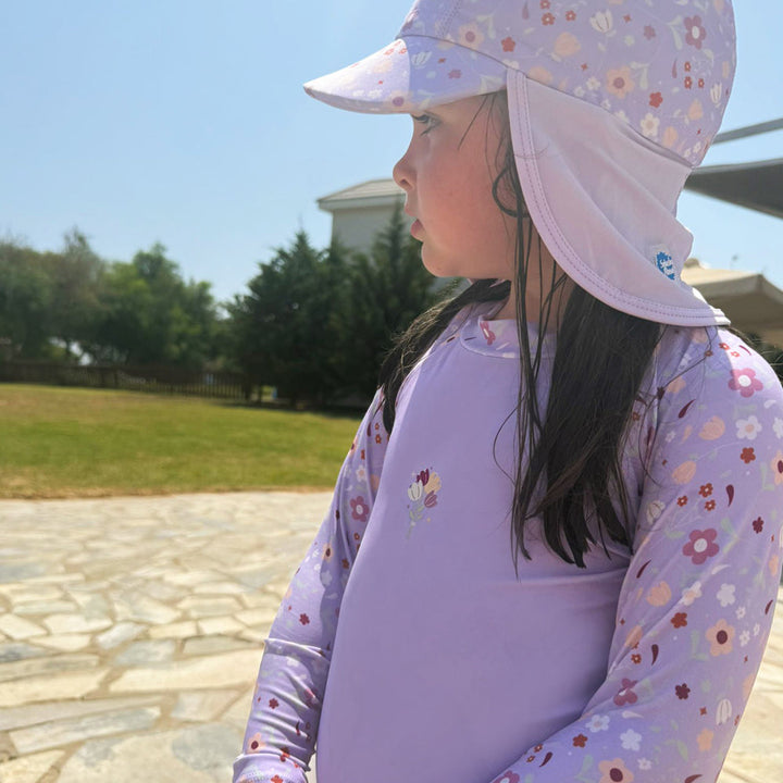 Little one wearing protective long sleeve rash top in lilac, and floral themed print on the chest and sleeves. They are also wearing matching Legionnaire Hat.