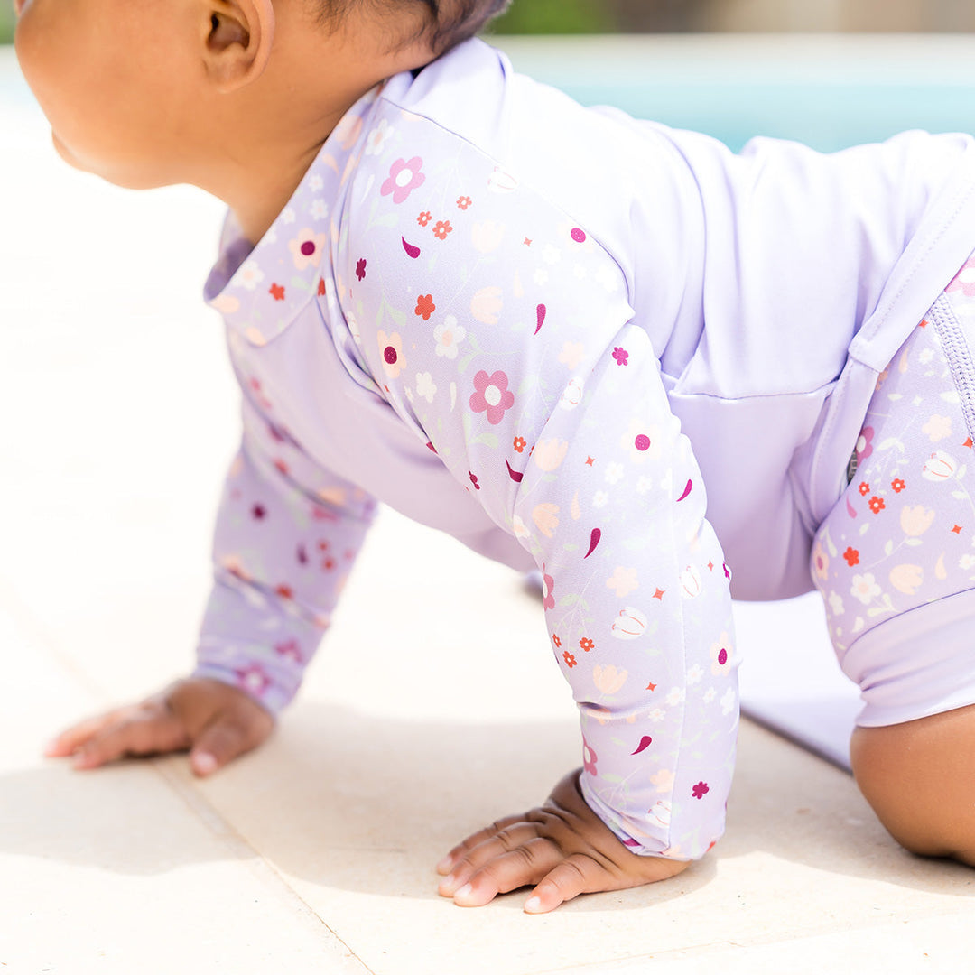 Close up of little one wearing protective long sleeve rash top in lilac, and floral themed print on the chest and sleeves. They are also wearing matching Happy Nappy.