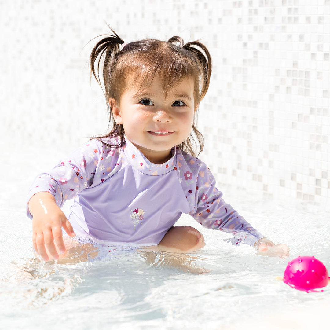 Little one wearing protective long sleeve rash top in lilac, and floral themed print on the chest and sleeves, in the pool.