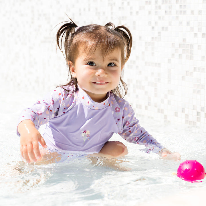 Little one wearing protective long sleeve rash top in lilac, and floral themed print on the chest and sleeves, in the pool.