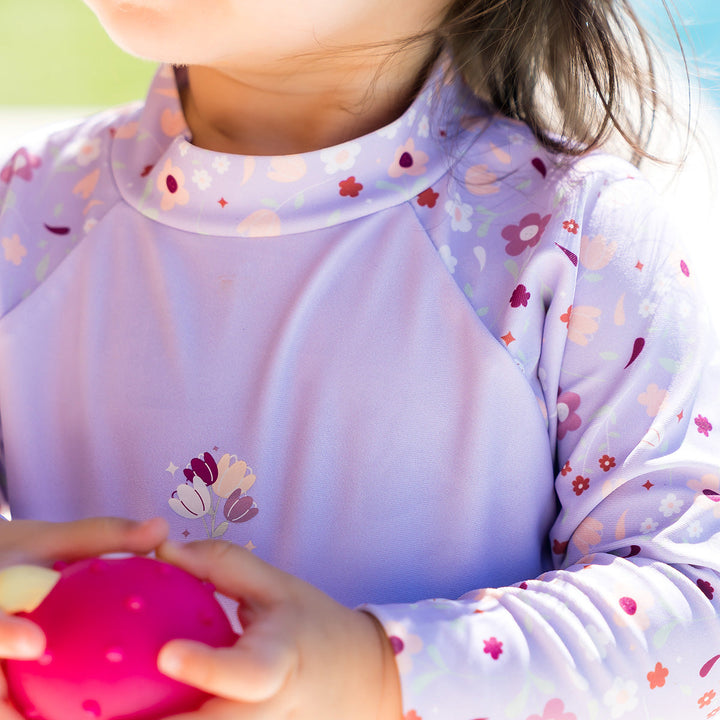 Close up of little one wearing protective long sleeve rash top in lilac, and floral themed print on the chest and sleeves..