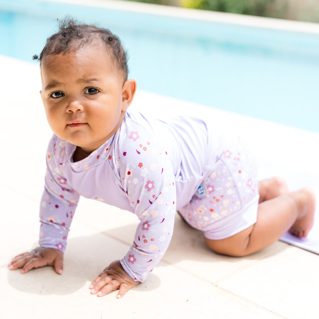 Little one wearing protective long sleeve rash top in lilac, and floral themed print on the chest and sleeves. They are also wearing matching Happy Nappy.