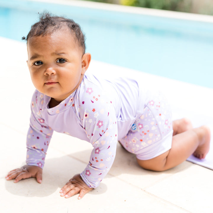 Little one wearing protective long sleeve rash top in lilac, and floral themed print on the chest and sleeves. They are also wearing matching Happy Nappy.