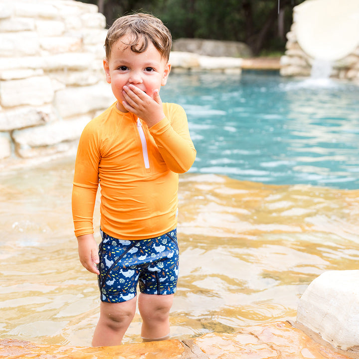 Little one wearing UV protective long sleeve rash top in neon orange. They are also wearing blue jammers.